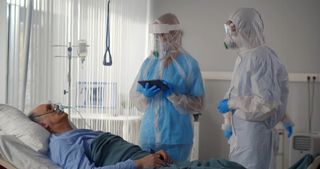 Doctors wearing protective uniform with gloves holding digital tablet standing near sick man in ward
