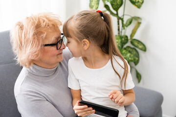 Happy grandmother and granddaughter using phone together, sitting on cozy sofa at home, browsing mobile device apps, grandma with grandchild playing game, looking at screen, having fun