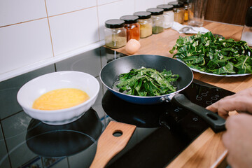 Preparing eggs with spinach for breakfast. Overhead view of black kitchen stove with frying pan.