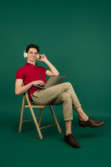 Typing text, listening to music. Caucasian handsome man's portrait on green studio background with copyspace. Male model with laptop. Concept of human emotions, facial expression, sales, ad, fashion.