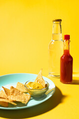 Nachos chips with cheese sauce on blue plate and soda bottle and hot sauce on yellow background. Food photo, vertical