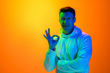 Nice sign. Caucasian man's portrait isolated on yellow studio background in mixed neon light. Handsome male model. Concept of human emotions, facial expression, sales, ad, fashion. Copyspace.