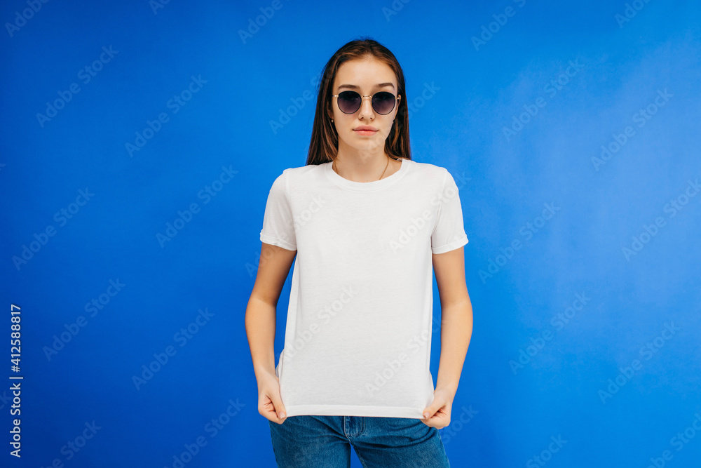 Poster Stylish girl in glasses wearing white t-shirt posing in studio on blue background