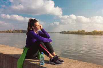 Young sweaty active fitness athletic sporty woman taking a break after outdoor exercising of running and jogging listening the music on her smartphone and enjoy the sunrise in the beautiful morning