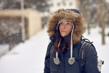 Middle-aged woman with backpack in winter snowfall wearing a fur trimmed warm jacket standing in an urban street looking up as she watches the snowflakes falling in a concept of the seasons
