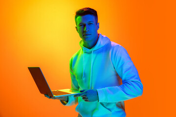 Scrolling laptop. Caucasian man's portrait isolated on yellow studio background in mixed neon light. Handsome male model. Concept of human emotions, facial expression, sales, ad, fashion. Copyspace.