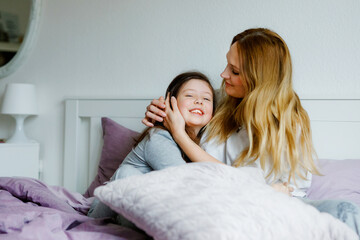 Lovely young mother and cute school kid girl cuddling together in bed in morning. Happy family of young single woman and beautiful daughter, indoors, getting ready and morning routine.