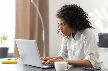 Young African American businesswoman with Afro hairstyle sitting at the desk, checking the stock trade market data online, looking at laptop screen, cannot believe her eyes, unbelievable news concept