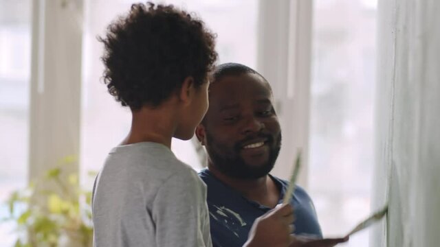 Happy African American father and cute little son smiling, playing and chatting while painting wall together during home renovation