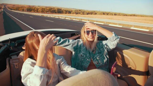 Women in sunglasses take selfies while posing in a convertible, happy and laughing. Two women on the back seat of a convertible driving on an empty highway, happy emotions of happiness