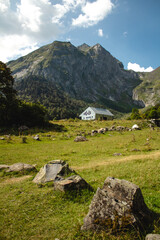 Fototapeta na wymiar Beautiful Wooden Shelter on Bright Green Grass with Trees Surrounded of Rocks, High Mountains and Peaks in a Sunny Cloudy Summer Day