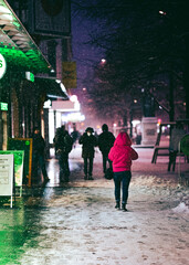 A girl with a purple jacket is going down a snowy shop window light illuminated street