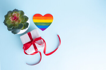 Gift box with a red ribbon, a home flower in a pot and a paper rainbow heart on a gentle blue background. LGBT congratulations concept