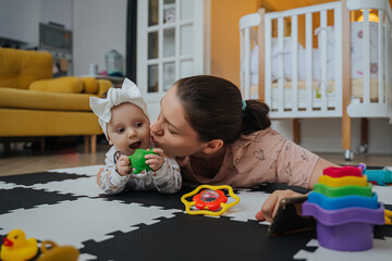 Young  mom kisses daughter lying on leisure rug. Happy  mom lying on carpet with adorable infant...