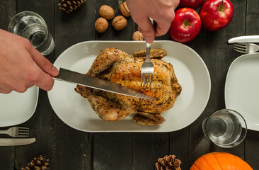 Male hands carving delicious homemade roasted chicken or turkey. Thanksgiving or Christmas flat lay composition on wooden table, decorated with apples, walnuts, cones and pumpkin.