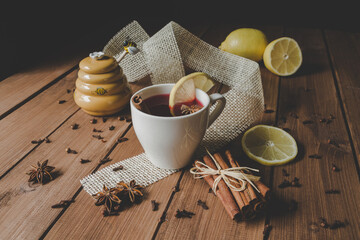 Warming cranberry tea on wooden table, with lemons, cinnamon sticks, star anise, cloves and yellow ceramic bee honey jar with honey dripping from a wooden honey dipper spoon.
