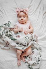 Astonished infant in trendy clothes and bow lying on soft bed under coverlet with floral ornament. Stylish baby with muslin swaddle blanket  on bed - obrazy, fototapety, plakaty