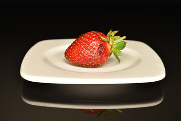 One berry of a bright red ripe organic delicious strawberry on a ceramic white saucer. The background is black.