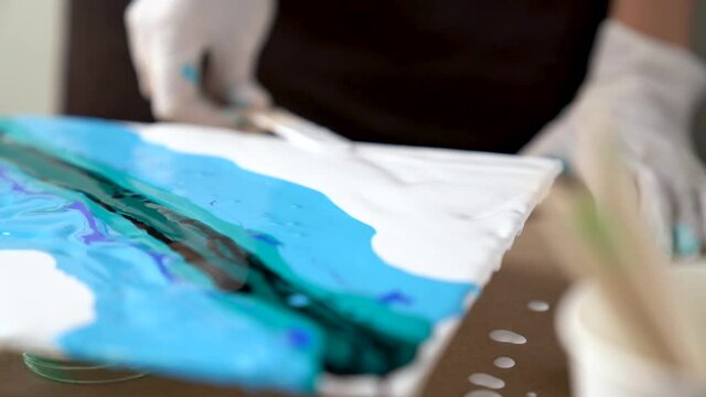 Closeup artist is smearing color with wooden stick across canvas. Creation of ornament on blue aquamarine picture. Process of drawing, making abstract liquid acrylic painting in workshop. Fluid art.