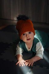 Funny baby on puzzle mat in dark room. Cute baby in trendy knitted hat lying on belly and playing on floor in dark room