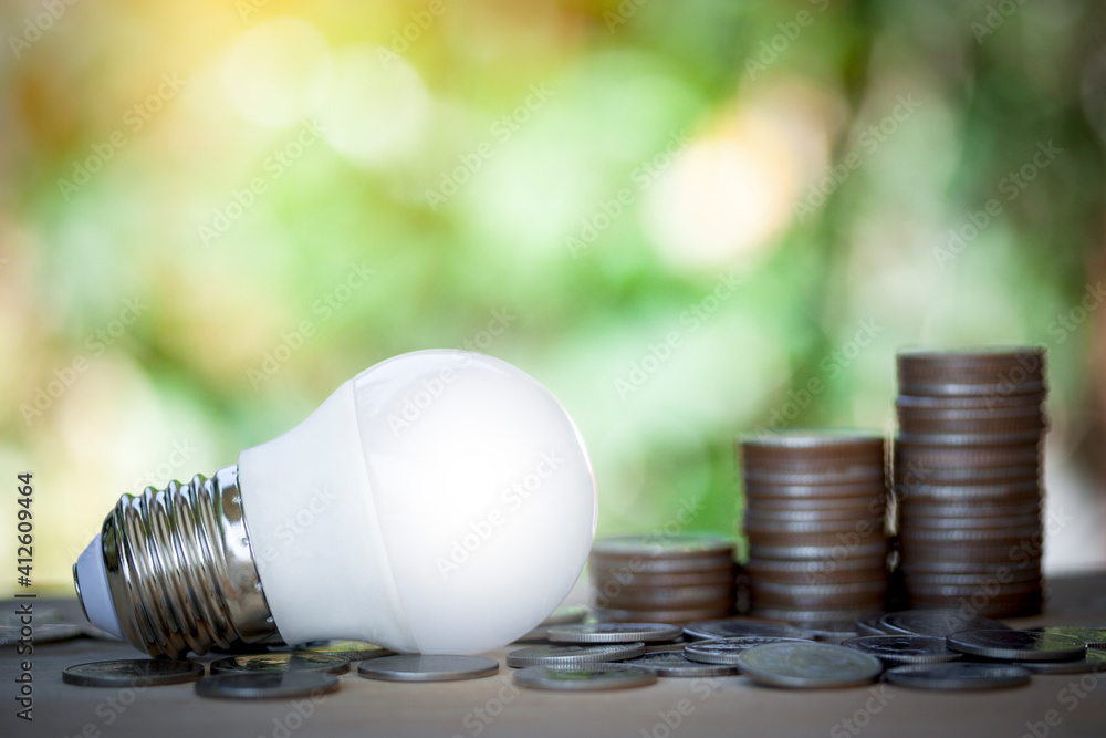 Poster led bulb on a lot of coins stack on a wooden table with light bokeh background saving energy concept