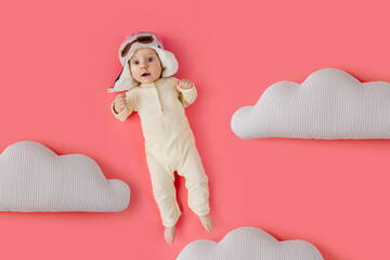 Top view of cute infant wearing pilot hat lying on pink background among soft toy clouds pretending...