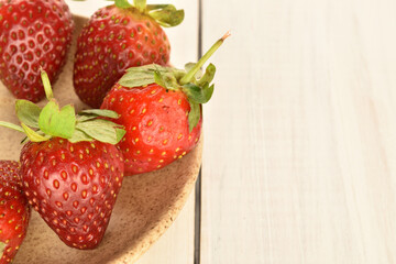 Several berries of bright red ripe organic delicious strawberries in a ceramic saucer on a wooden tabletop painted in white.