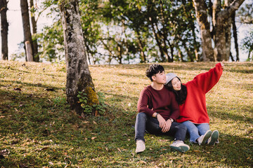 Naklejka na ściany i meble Portrait sweet couple of teenage sitting on hill, asian man with maroon sweater hug or cuddle a young woman with red sweater. Romantic mood and sweet emotional relationship in summer with copy space