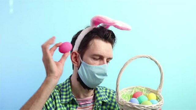 man in bunny ears and face mask  with wicker basket with eggs shakes an egg happy easter during coronavirus 