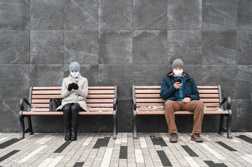 Young caucasian man and woman keep social distance looking at their smartphones and wearing protective face masks. They sit on benches against backdrop of stone wall in outdoors. Concept new normality