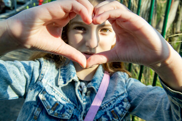 teenage girl with long hair looks through folded fingers in the shape of a heart on a sunny day