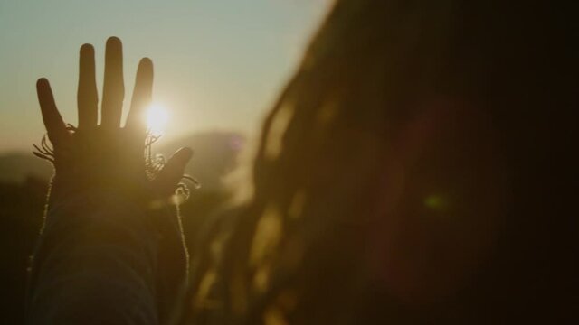 Woman with dreadlocks looking at sun through her fingers at sunset and dreaming. Close-up of hand at sunrise. Freedom concept. 
