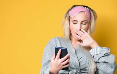 Young woman yawns and looking to smart phone. Sleepy girl yawning, model wearing nightie - night dress, isolated on yellow background