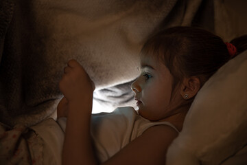 Cute baby girl uses phone in bed at night instead of sleeping close up.