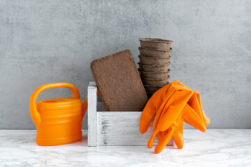 A white box with peat pots, coconut briquettes, orange gloves and an orange watering can on a gray concrete background