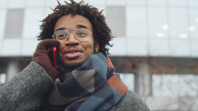 Chest up arc shot of young Afro-American man standing outdoors on winter day and speaking on mobile phone