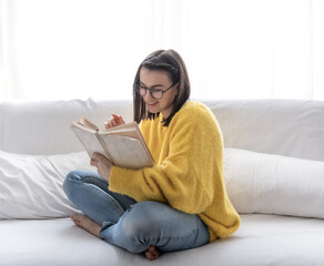 A smiling girl in glasses and a yellow sweater is sitting on the sofa with a book.
