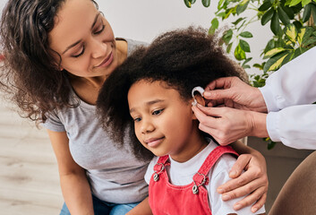 Hearing treatment for a child. African American girl with her mother during installation hearing aid by her audiologist - Powered by Adobe