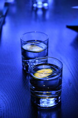 Two glasses of still water with cucumber pieces to refresh. Vibrant blue neon lighting to summer cocktails on wooden background colored in shades of blue due to neon light