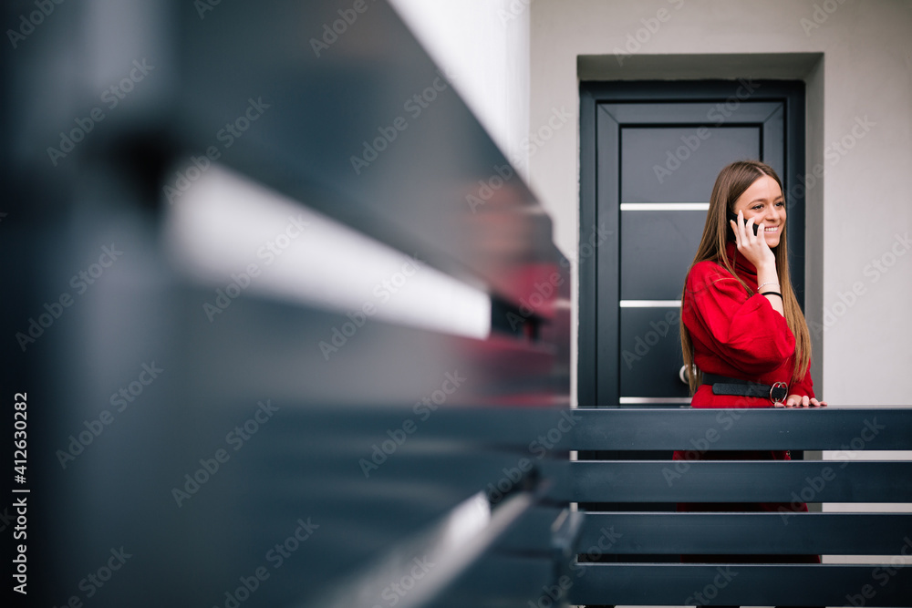 Wall mural Young woman in a red dress having a telephone conversation at the door of the house. Teenager talking on the phone. Selective focus