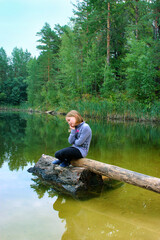 Little sad girl sitting on the log by the lake. Melancholy child by forest lake