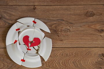 A white plate is broken on the wooden floor. We tried to collect the pieces of broken dishes and glue them with tape. The red heart is split and lies on a broken dish, crockery shards.
