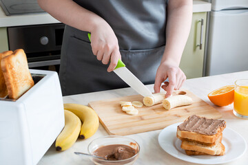 Woman cuts banana for toast for breakfast. European breakfast with toast, banana and juice.