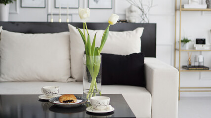 Sofa in a modern interior. On the table are a bouquet of tulips, coffee cups and a sweet bun. Black and white interior and sunny mood.