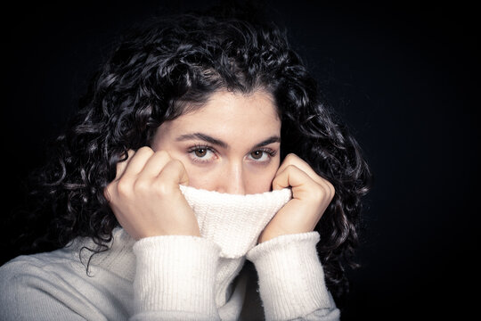 Girl covering her mouth with the neck of her sweater for fear of being infected with visrus, isolated on black background, concept of phobia and alarmism