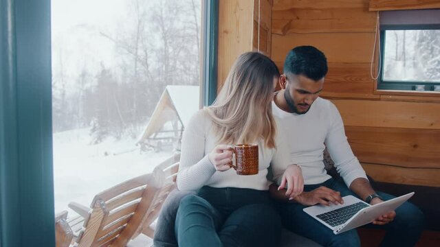 Happy young couple enjoying winter vacation. Browsing social media on laptop and drinking hot drink. High quality 4k footage