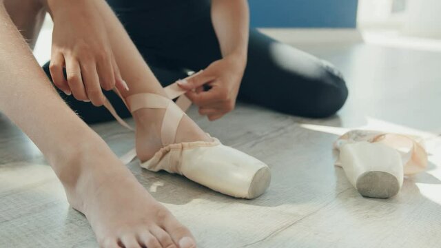 Close up of young beautiful caring ballet teacher shows ittle girl how to tie light pointe shoes on her leg.