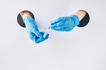 Coronavirus prevention vaccine in protective medical blue gloves in hand through white paper hole. Vaccination shots to prevent infectious diseases.