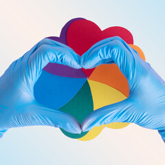 Heart shape from hands in blue protective gloves over a rainbow flower made of multicolored hearts on a pink background, close-up, square frame. LGBT concept. Safe love concept