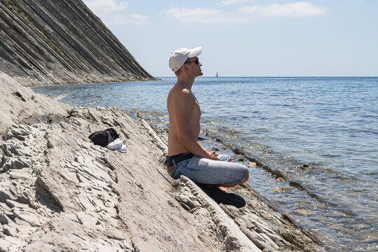 Summer sea landscape. The guy sits on a stone wild beach and looks into the distance. On the horizon, clear blue sky and steep cliffs. The resort city of Gelendzhik. Russia, Black Sea coast